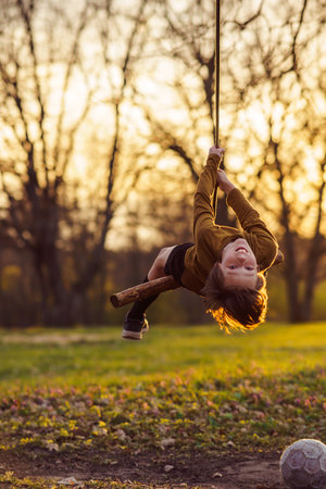 Smiling teenage boy is hanging upside down on homemade swing in nature, in park, in village in rays of sunset. Outdoor games and entertainment. Happy childhood. Summer in country. Child protection.の写真素材