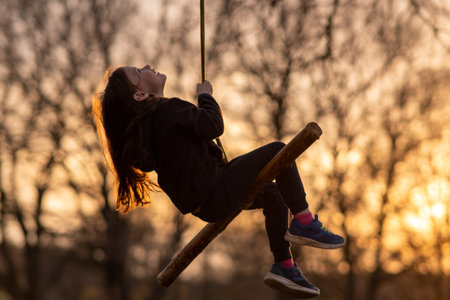 Portrait of happy little girl who swings on homemade swing in nature, in park, in village, in children's camp against background of trees. Outdoor games and entertainment. Child protection.の写真素材