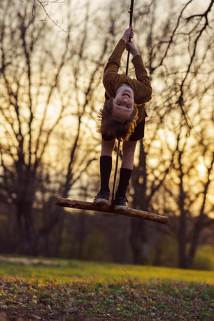 Happy teenage boy hanging upside down on homemade swing in nature, in park, in village in sunset rays in evening. Positive emotions. Child protection. Child safety. Autumn portrait.の写真素材