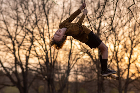 Happy teenage boy flies high up on homemade swing in nature, in park, in village against background of autumn trees in colors of sunset. Positive emotions. Child protection. Child safety. Recreation.の写真素材