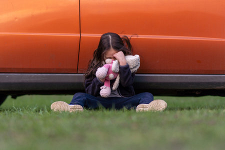 Little lonely girl is crying, hiding her face in toy, sitting on grass with her back pressed against car. Children's tears. Child is upset and offended. Children's problems. Inner world of child.の写真素材
