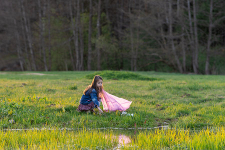 Charming little girl with long hair in denim jacket collects garbage in nature, in park, in meadow on summer day. Environmental problems. We take care of nature. Saving planet.の写真素材