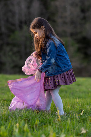 Vertical portrait of charming little girl with long hair in denim jacket collecting garbage in nature, in meadow on summer day. Environmental problems. We take care of nature. Saving planet.の写真素材