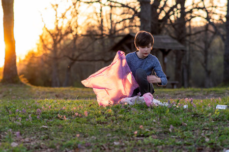 Children's volunteer activity. Teenage boy collects garbage in large pink bag in nature, in meadow on autumn evening at sunset. Environmental problems. We take care of nature. Saving planet.の写真素材