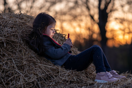 Beautiful portrait of charming little girl lying on haystack playing with her phone against background of setting sun. Happy childhood. Summer in country. Time to dream. Inner world of child.の写真素材