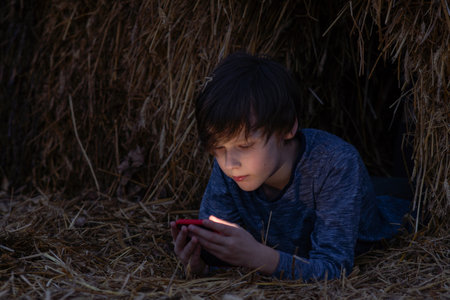 Boy lies in haystack on summer night and reads something exciting from phone screen. Happy childhood. Summer holidays. Summer in country. Time to dream. Inner world of child. Kids are outside.の写真素材