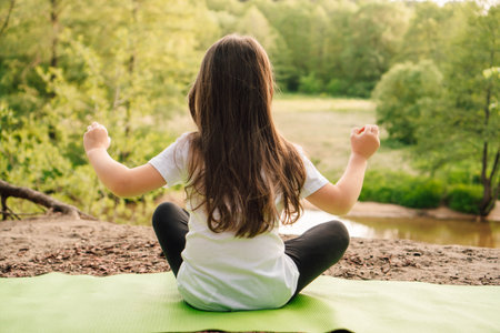 Rear view of little girl with brown long hair sitting on green sporty rug and practicing yoga in front of river. Active female sportsman in white t-shirt and black leggings meditating in nature.の写真素材