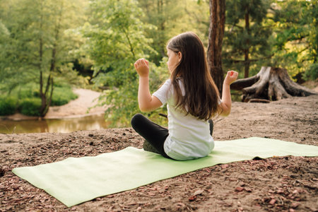 Rear view of female sporty child peacefully sitting on green mat in yoga pose with raised arms. Little girl with long brown hair closed eyes and doing sports on hill in front of river in forest.の写真素材