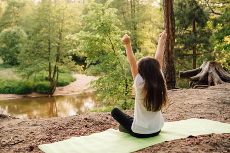 Rear view of little girl with brown and long hair comfortable sitting on green sporty mat and rising arms while enjoying nature. Active female child spending time resting on hill in front of river.の写真素材