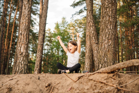 Adorable and happy female kid with open mouth sitting on ground near trees in forest with raised arms. Little sporty girl with dark hair enjoying spending active time in nature.の写真素材