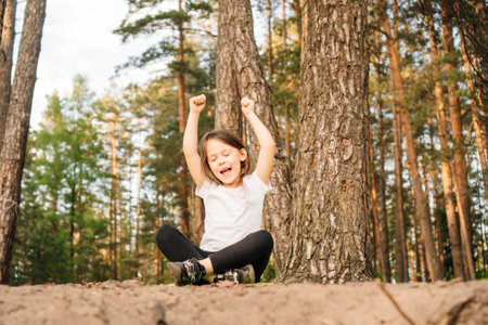 Delightful little girl in white t-shirt and black leggings sitting on ground among trees with closed eyes and raised arms. Happy active child with dark and long hair enjoying time outdoor.の写真素材