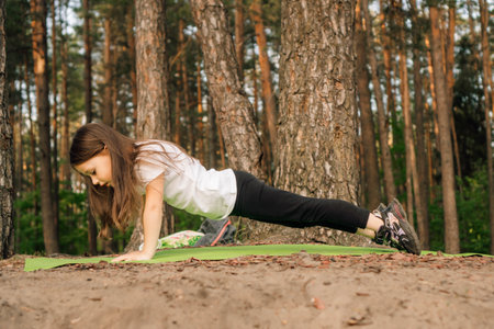 Strong little female sportsman with dark hair standing on plank exercise on green sporty mat in nature near trees. Focused athletic girl in white t-shirt and black leggings training outdoor.の写真素材
