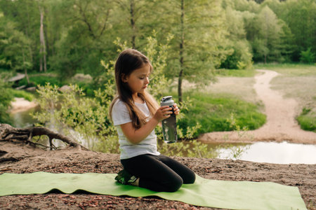 Charming little lady in white t-shirt and black leggings sitting on green sporty mat outdoor and holding bottle of water in hands. Athletic child looking at flask, resting on hill in front of river.の写真素材