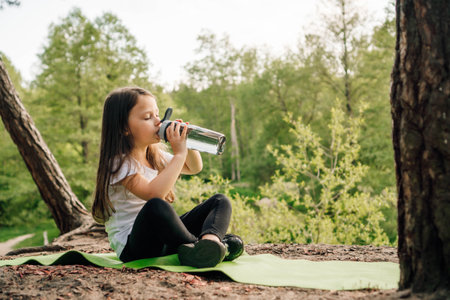 Adorable little girl with dark long hair sitting on green sporty rug in nature and drinking water from bottle. Junior female sportsman resting after workout outdoor.の写真素材