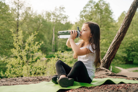 Adorable little girl with dark long hair sitting on green sporty rug in nature and drinking water from bottle. Junior female sportsman resting after workout outdoor.の写真素材
