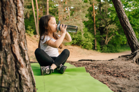 Adorable little girl with dark long hair sitting on green sporty rug in nature and drinking water from bottle. Junior female sportsman resting after workout outdoor.の写真素材