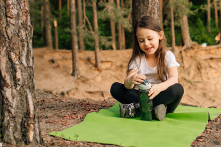 Delightful female kid sitting on green rug outdoor and opening bottle of water that holding in hands. Smiling girl with brown long hair looking at flask and spending time in fresh air.の写真素材