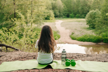 Rear view of little female sportsman with brown long hair sitting on green mat on sandy hill in front of lake and enjoying nature. Small dumbbells and bottle of water lying near child.の写真素材