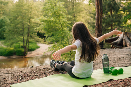Rear view of female kid with arms wide open sitting on green mat in nature with bottle of water and small dumbbells near. Little athlete with dark hair enjoying time on sandy hill in front of river.の写真素材