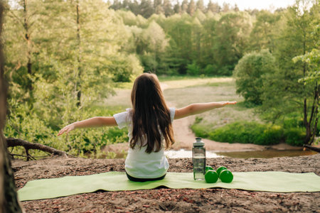 Rear view of brunette little girl with arms wide open sitting on green sporty rug on sandy ground outdoor. Child resting on hill in front of river, training equipment and bottle lying near athlete.の写真素材