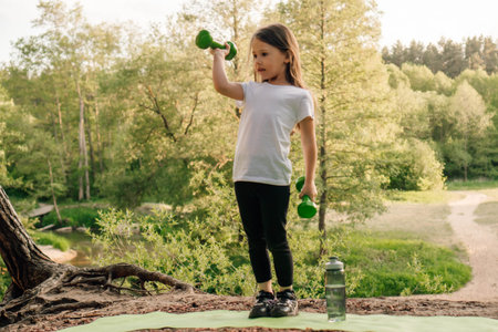 Dynamic junior female sportsman standing on green sporty mat outdoor and working out with small dumbbells in hands. Little girl with dark hair lifting weights and actively training in nature.の写真素材