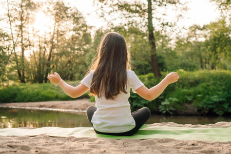 Back view of someone sitting on green sporty mat on sandy beach in yoga pose with arms wide open. Person with long hair wearing comfortable clothes doing sports outdoors next to lake.の写真素材