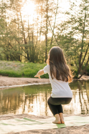 Rear view of junior sporty girl with long flowing hair doing squats in front of lake on sandy plage. Active child in white t-shirt and black leggings exercising in nature at sunset. Vertical.の写真素材
