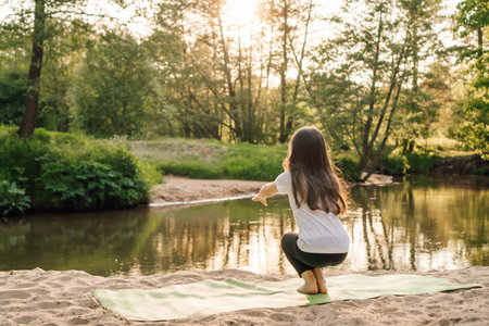 Rear view of little girl with stretched arms forward doing narrow squats on green mat on sandy beach near lake at sunset. Junior athlete with long hair actively spending time doing sports in nature.の写真素材