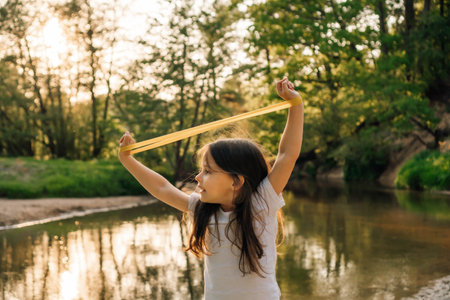 Adorable little girl raising arms with mini band on wrists above head and stretching sporty equipment while standing close to lake in forest. Junior athlete with dark hair training at sunset.の写真素材