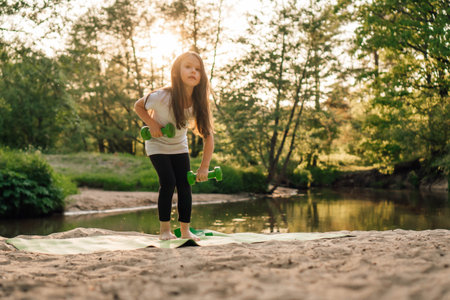Adorable junior female sportsman lifting green small dumbbells from ground standing on sporty mat on sandy beach near river. Little girl with dark long hair working out outdoor in forest.の写真素材