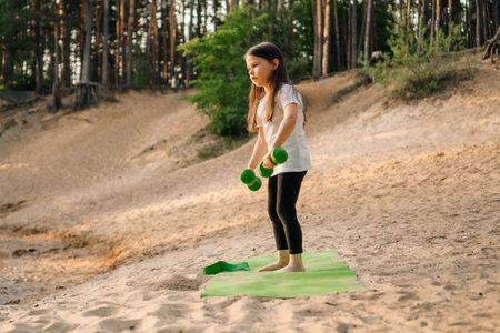 Energetic little lady in white t-shirt and black leggings doing workout with small dumbbells in hands standing on green sporty rug outdoor. Junior athlete training on sandy beach in forest.の写真素材