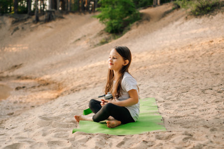 Adorable little girl with brown long hair sitting on green mat outdoor with bottle of water in hand. Female kid smiling while resting on sporty rug on sandy beach in forest.の写真素材