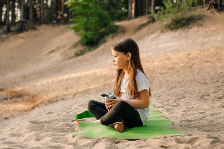 Charming little lady comfortable sitting on green sporty mat on sandy beach and holding bottle of water in hands. Brunette child looking at flask with liquid, small dumbbells lying near kid.の写真素材