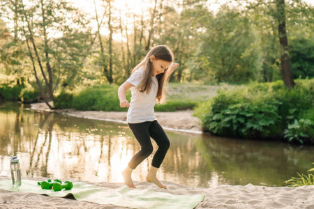 Delightful little athlete with dark long hair happily jumping on green mat on sandy beach near river. Smiling girl in white t-shirt and black leggings doing sports outdoors.の写真素材