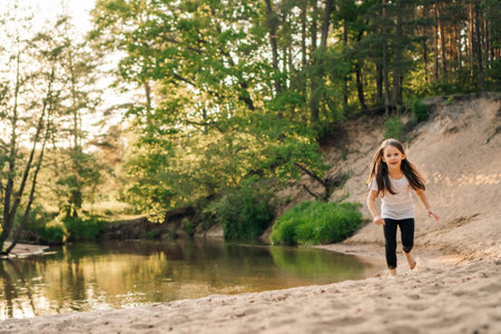 Happy, energetic girl in white t-shirt and black leggings running on sandy beach close to river. Little athlete with dark long hair doing sports and having fun in forest near water.の写真素材