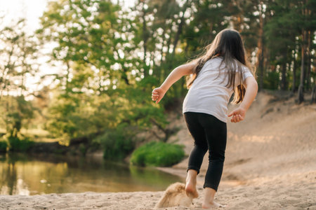 Rear view of little brunette with long hair running on sandy beach near water. Active girl wearing white t-shirt and black leggings jogging in nature close to river.の写真素材
