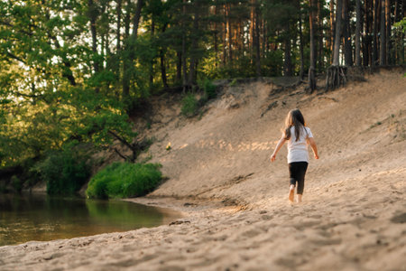 Rear view of little girl with dark, long and flowing hair running on sandy beach near river in forest. Female child in white t-shirt and black leggings actively spending time in nature.の写真素材
