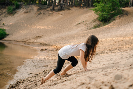 Energetic girl with brown long hair climbing up small hill on sandy beach near river. Little athlete in white t-shirt and black pants actively spending time outdoors walking close to water in forest.の写真素材