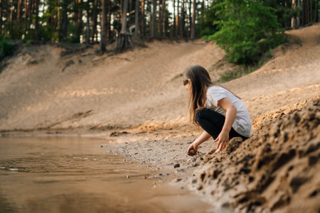 Curious little girl with brown flowing hair squatting down on sand near river in forest and going to take grit. Water reflect child in white t-shirt and black trousers.の写真素材