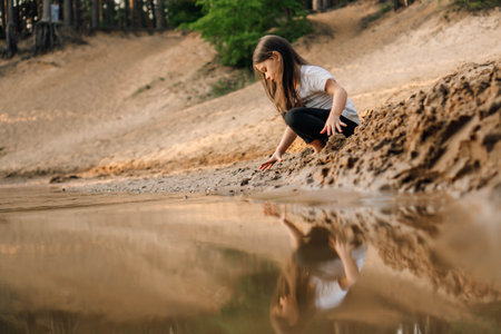 Curious little girl with brown flowing hair squatting down on sand near river in forest and going to take grit. Water reflect child in white t-shirt and black trousers.の写真素材