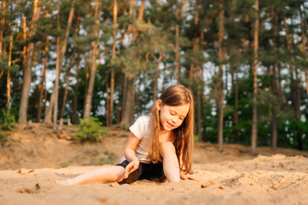 Adorable little girl with dark and long hair comfortable sitting on sand near forest. Female child wearing white t-shirt and black shorts playing with grit and having fun in nature.の写真素材