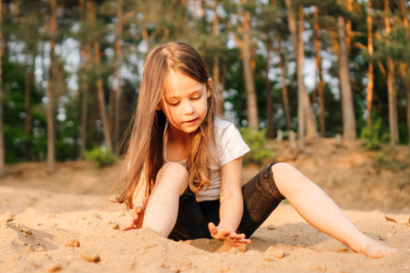 Adorable little girl with dark and long hair comfortable sitting on sand near forest. Female child wearing white t-shirt and black shorts playing with grit and having fun in nature.の写真素材