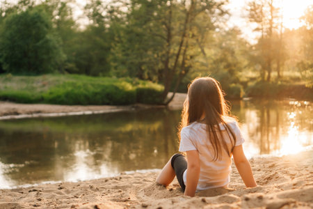 Adorable little girl with dark and long hair comfortable sitting on sand near forest. Female child wearing white t-shirt and black shorts playing with grit and having fun in nature.の写真素材