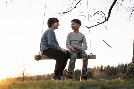 Friendship. Two teenagers, friends, brothers, classmates are sitting on swing against light gray sky at sunset on autumn evening and talking. Communication. Happy childhood. Free time.の写真素材