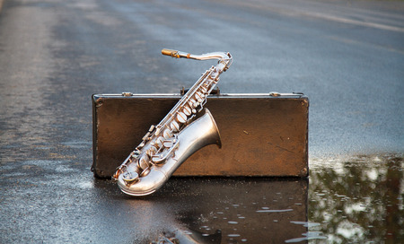 Silver saxophone based on a suitcase standing on an empty, asphalt, wet roadの写真素材