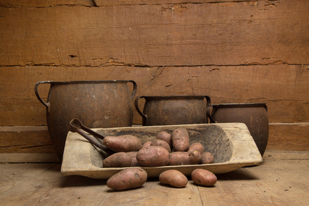 Old cast-iron pots, potatoes in a wooden bowl standing on a wooden floor against the background of a wooden wallの写真素材