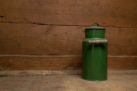 An old metal, green varnished milk can on a wooden floor against the background of a plank wall.の写真素材