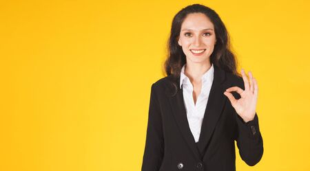 Beautiful of young woman Caucasian portrait. Smiling businesswoman black long curly hair showing finger ok gesture and happy looking at the camera, isolated on yellow background with copy space.の写真素材