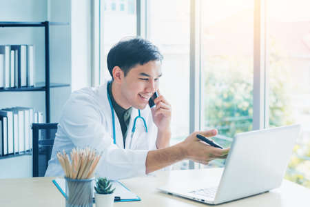 Portrait handsome young medical Asian doctor smiling has stethoscope talking smartphone provide patient consultation check symptom disease on laptop notebook on desk office clinic. Healthcare concept.の写真素材