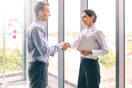 Handshake successful business together, portrait of young caucasian businessman and businesswoman happy smiling standing near window in office. Partnership finance teamwork contract business concept.の写真素材